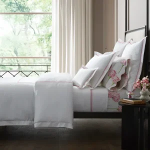 A stylish bedroom with lots of light featuring a bed with amara triple pearl white bedding, with beaded pink and red pearl-like embroidery strands, and floral accents from the Charlotte collection, set against a backdrop of greenery outside.