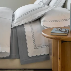 A cozy bed featuring white scalloped bedding with grey trim, complemented by a wooden nightstand holding a book and glasses. Featuring Classic Chain Scallop bedding by Matouk. Available at Linen Society.