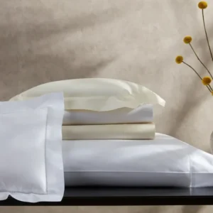 A stack of soft, white and ivory sheets and pillowcases with a delicate hemstitch on a dark wooden surface, accompanied by a vase of yellow flowers. Featuring Sierra Hemstitch bedding by Matouk, available at Linen Society.