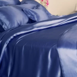 A bed features Kumi Kookoon’s Classic Silk fitted sheet in blue charmeuse, with matching pillowcases and the blanket partially pulled back.